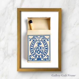 May include: A framed print featuring a matchbox with a blue and white design. The matchbox has a horseshoe and clover graphic with the words "LUCKY YOU". A single match with a black tip is visible. The frame is gold.