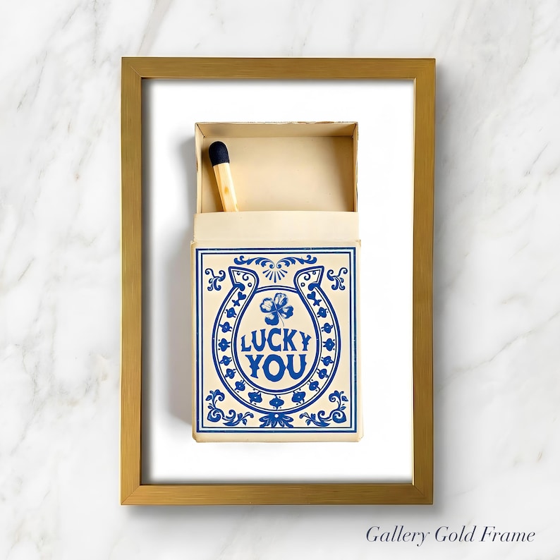 May include: A framed print featuring a matchbox with a blue and white design. The matchbox has a horseshoe and clover graphic with the words "LUCKY YOU". A single match with a black tip is visible. The frame is gold.