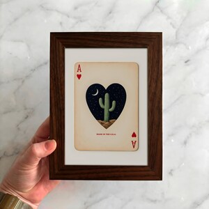 May include: Framed playing card art featuring the Ace of Hearts. The card displays a heart-shaped design with a cactus under a crescent moon and stars. The text "MADE IN THE U.S.A." is visible. The frame is dark brown.
