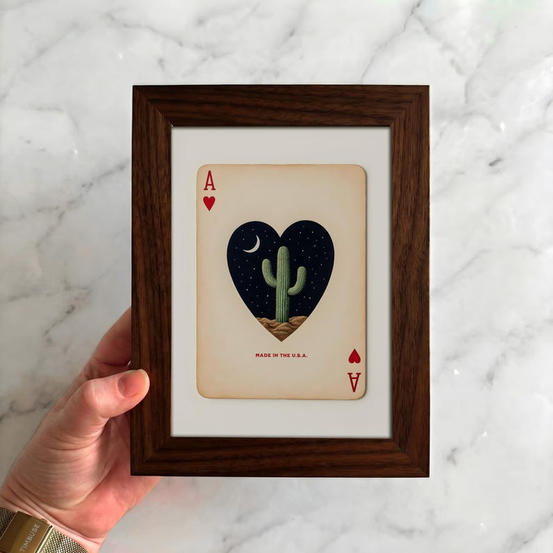 May include: Framed playing card art featuring the Ace of Hearts. The card displays a heart-shaped design with a cactus under a crescent moon and stars. The text "MADE IN THE U.S.A." is visible. The frame is dark brown.