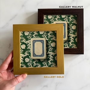 May include: Two framed art pieces. One frame is gold, the other is dark brown. Each frame contains a small, rectangular, off-white object with a blue border, set against a green floral patterned background. The text "GALLERY GOLD" and "GALLERY WALNUT" are visible.