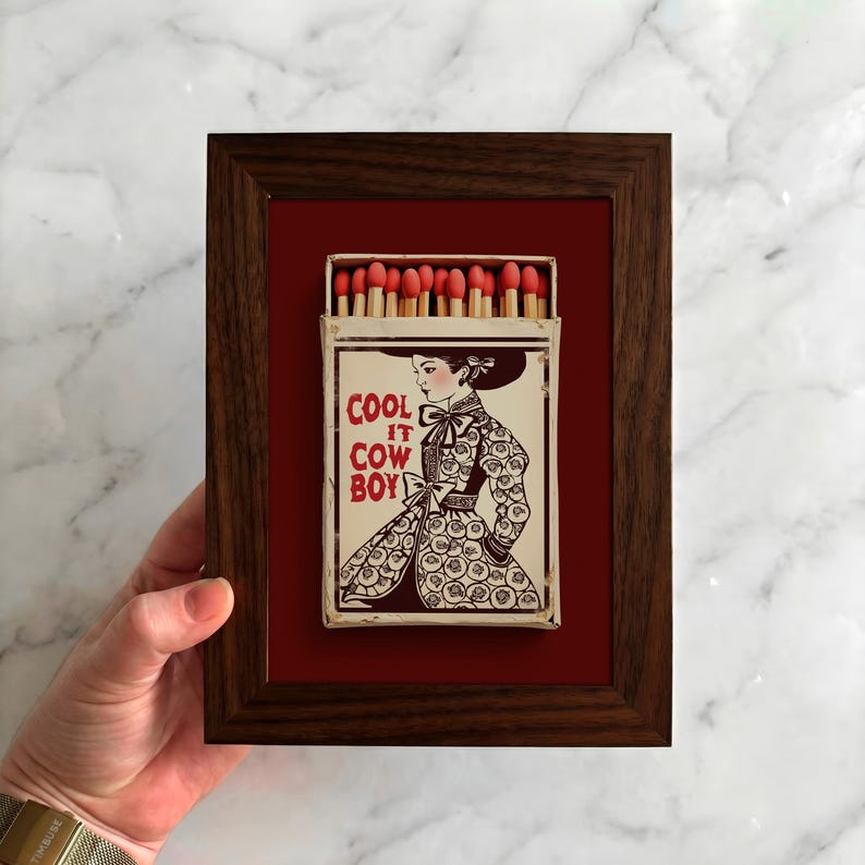 Op de afbeelding: Een ingelijst kunstwerk met een luciferdoosje-illustratie. Het luciferdoosje, tegen een rode achtergrond, is ingesloten in een donkerbruine houten lijst. De illustratie toont een vrouw met de tekst "COOL IT COW BOY".