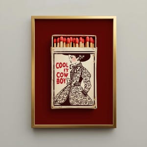 Vintage-inspired framed matchbox art featuring a classic cowgirl illustration and the phrase "Cool It Cowboy" on a weathered matchbox with red-tipped matches, set against a rich burgundy background in a gold frame. Americana western wall art with an heirloom, collected aesthetic.