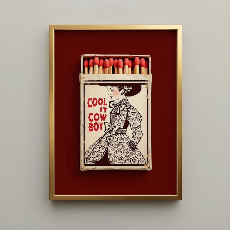 Vintage-inspired framed matchbox art featuring a classic cowgirl illustration and the phrase "Cool It Cowboy" on a weathered matchbox with red-tipped matches, set against a rich burgundy background in a gold frame. Americana western wall art with an heirloom, collected aesthetic.