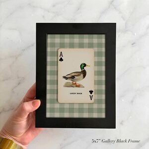 May include: A black 5x7 inch gallery frame containing a playing card with an Ace of Clubs symbol and a mallard duck illustration. The card features the text LUCKY DUCK and is set against a light green and white gingham patterned background.