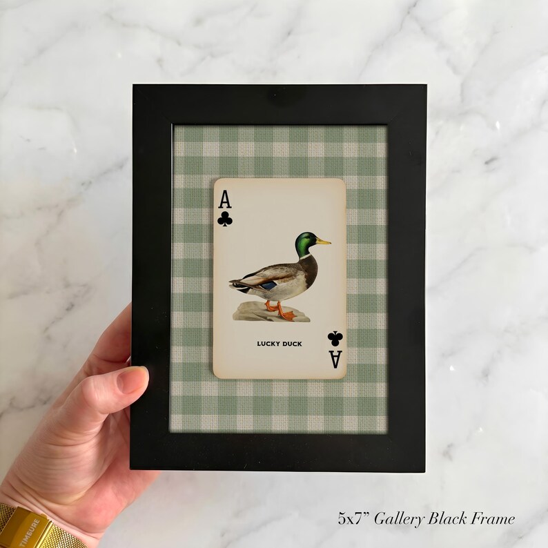 May include: A black 5x7 inch gallery frame containing a playing card with an Ace of Clubs symbol and a mallard duck illustration. The card features the text LUCKY DUCK and is set against a light green and white gingham patterned background.