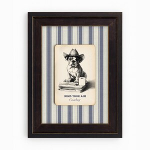 May include: Framed artwork featuring a bulldog wearing a cowboy hat. The black and white illustration is set against a blue and white striped background. The text "MIND YOUR AIM Cowboy" is printed below the dog.