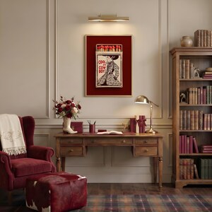Op de afbeelding: Een ingelijst kunstwerk met een lucifersdoosontwerp met de tekst "COOL IT COW BOY!" en een vrouw in een jurk. Het kunstwerk staat tegen een rode achtergrond en wordt tentoongesteld boven een houten bureau met een lamp en boeken.