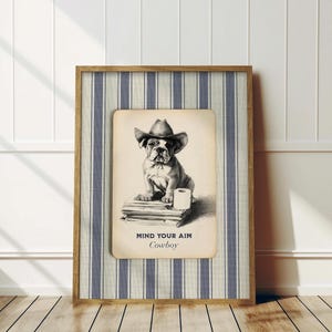 May include: A framed print featuring a bulldog wearing a cowboy hat. The black and white illustration shows the dog sitting on books with a roll of toilet paper. The text "MIND YOUR AIM Cowboy" is below the image. The frame has a striped background.