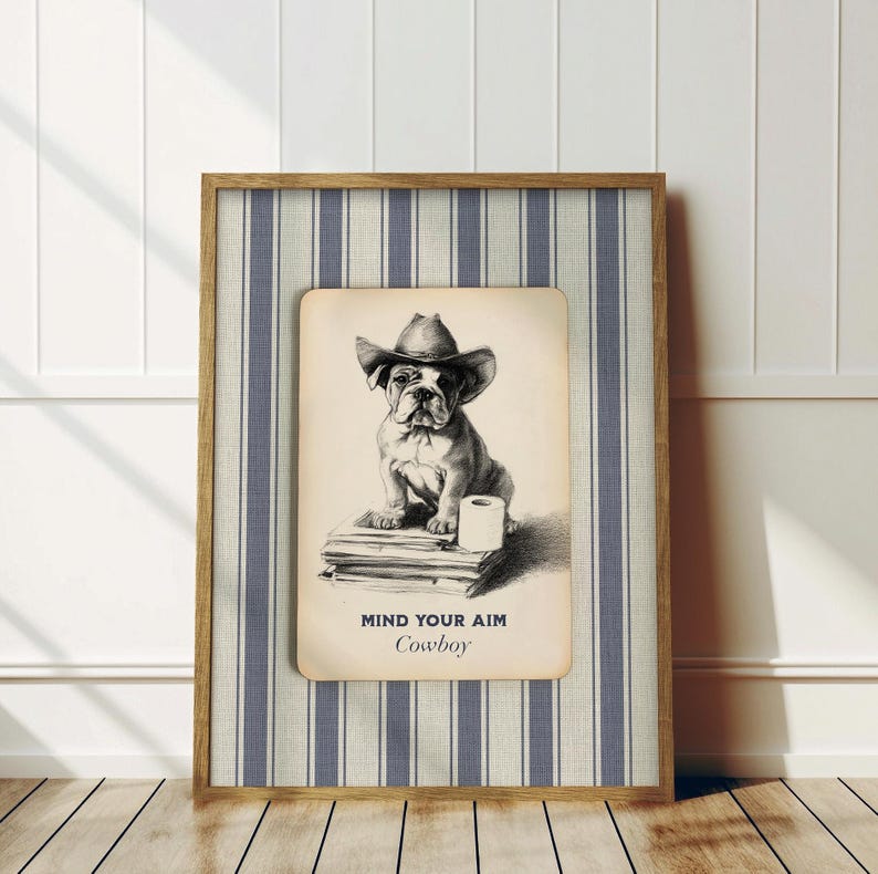 May include: A framed print featuring a bulldog wearing a cowboy hat. The black and white illustration shows the dog sitting on books with a roll of toilet paper. The text "MIND YOUR AIM Cowboy" is below the image. The frame has a striped background.