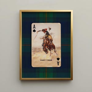 Vintage cowboy playing card wall art featuring an Ace of Spades with an expressive rodeo rider and the phrase "Easy Tiger," set against a deep green and navy tartan plaid background. Framed in a gold gallery frame as masculine Americana western decor.