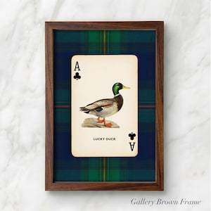 May include: Framed art print featuring a vintage playing card design. The card depicts a mallard duck illustration with the text "LUCKY DUCK" and the Ace of Clubs symbol. The frame is a dark brown color, and the background is a plaid pattern.