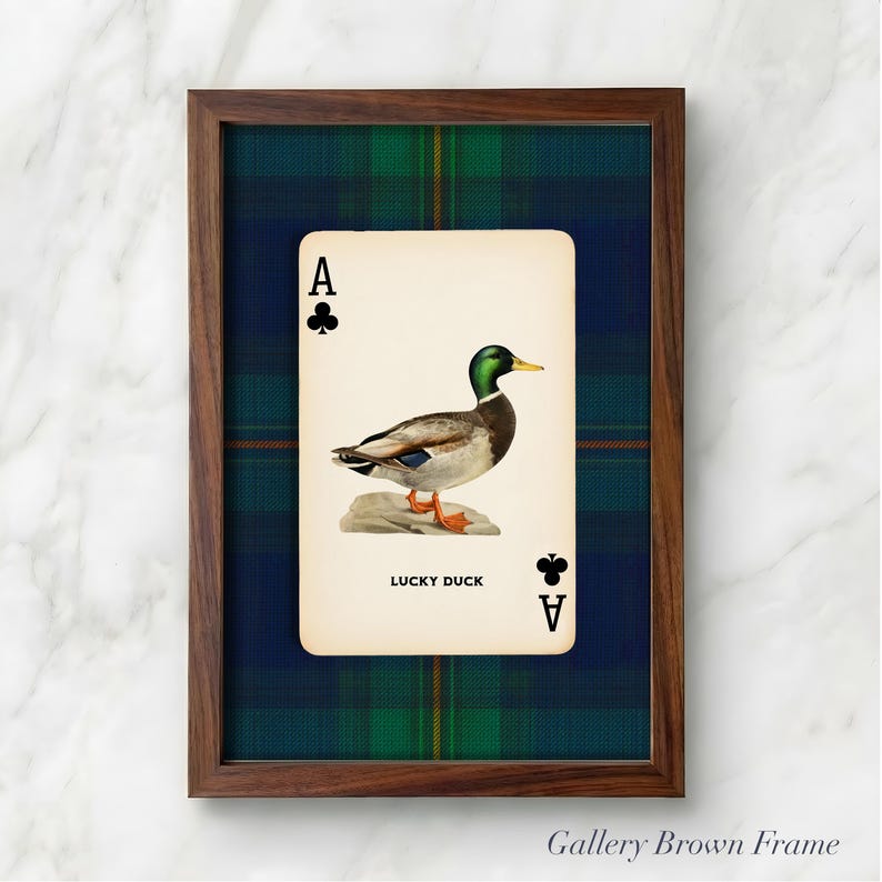 May include: Framed art print featuring a vintage playing card design. The card depicts a mallard duck illustration with the text "LUCKY DUCK" and the Ace of Clubs symbol. The frame is a dark brown color, and the background is a plaid pattern.