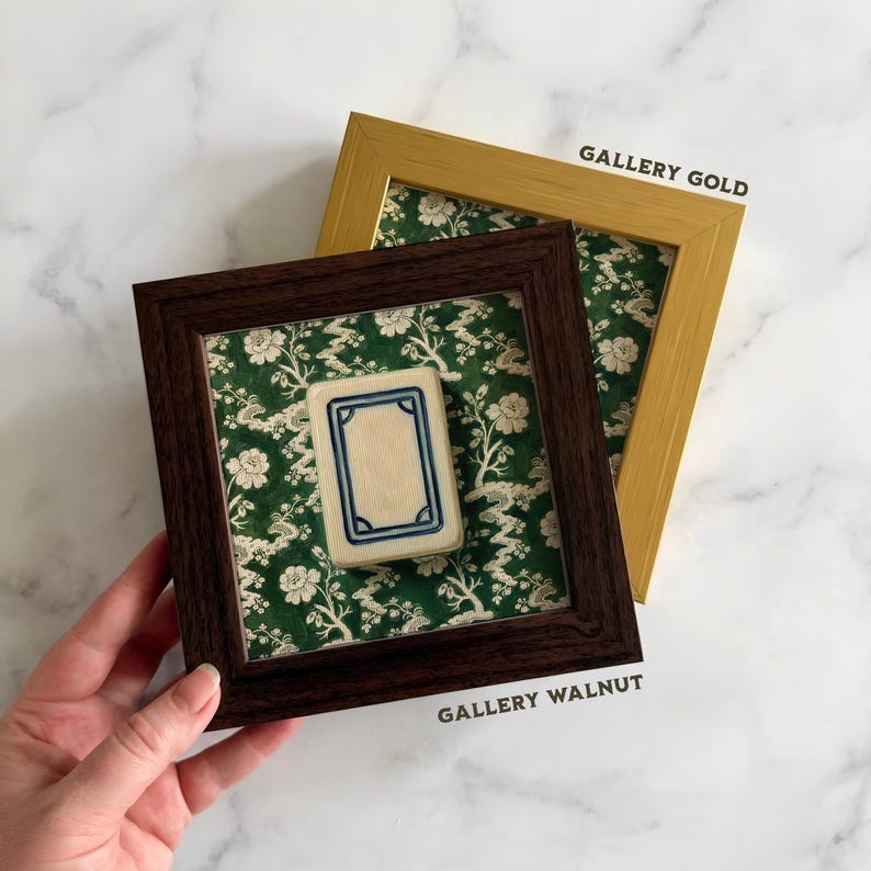 May include: A framed Mahjong tile with a cream-colored center and blue border, set against a green floral patterned background. The frame is dark brown, labeled "GALLERY WALNUT". A gold frame, labeled "GALLERY GOLD", is in the background.