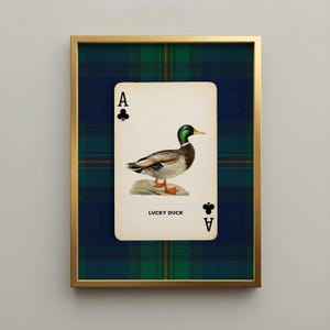 May include: A framed print featuring a playing card design with a mallard duck illustration. The card is labeled "Lucky Duck" and framed in gold, set against a plaid background. The card shows the Ace of Clubs.