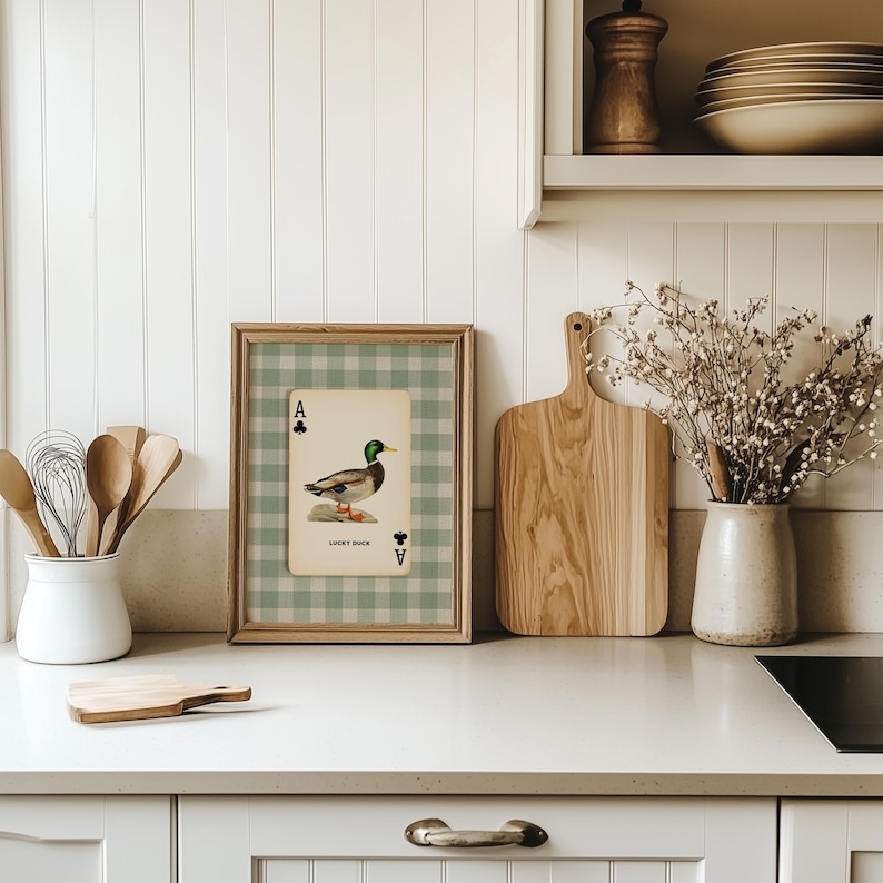 May include: A framed print featuring a playing card design with a duck illustration and the text "LUCKY DUCK." The card is set against a green and white checkered background. The frame is made of light-coloured wood. The print is displayed on a white countertop.