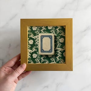 May include: A framed tile with a cream-colored center and a blue border, set against a green floral patterned background. The frame is gold-colored. The tile is displayed in a square wooden frame.