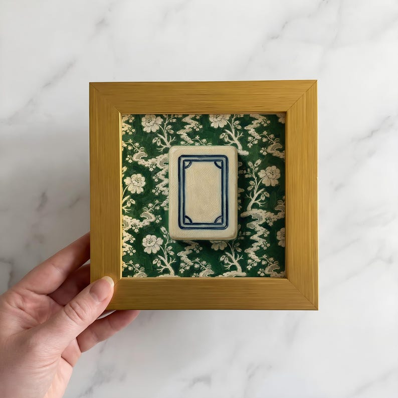 May include: A framed tile with a cream-colored center and a blue border, set against a green floral patterned background. The frame is gold-colored. The tile is displayed in a square wooden frame.