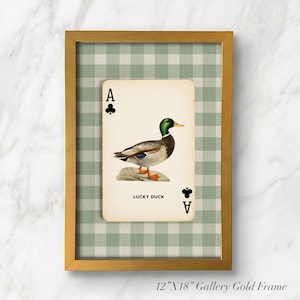 May include: A framed wall art piece featuring a playing card design with a mallard duck illustration, the text LUCKY DUCK, and club symbols. The card is set against a green and white gingham background inside a 12-inch by 18-inch gold frame.