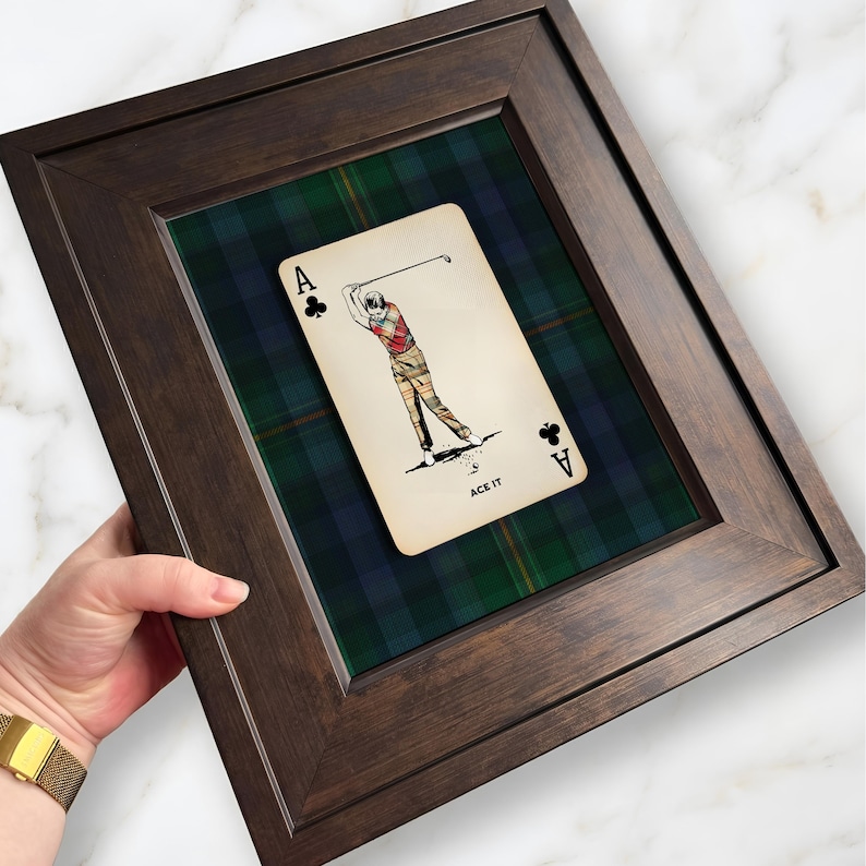 May include: Framed artwork featuring a playing card with a golf theme. The card depicts a golfer in action, set against a green and blue plaid background. The dark brown frame surrounds the artwork, with the text "ACE IT" at the bottom.