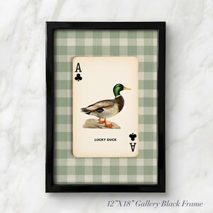 May include: A 12-inch by 18-inch black gallery frame containing a playing card print featuring an illustration of a mallard duck standing on a rock. The card displays the text LUCKY DUCK and club symbols. The background is a green and white gingham pattern.