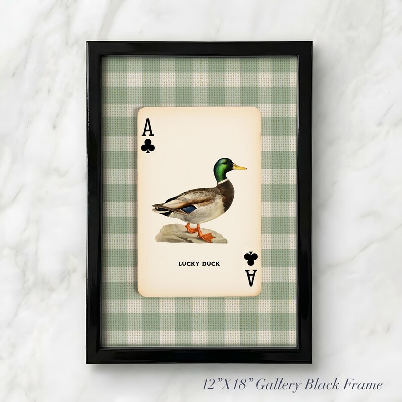 May include: A 12-inch by 18-inch black gallery frame containing a playing card print featuring an illustration of a mallard duck standing on a rock. The card displays the text LUCKY DUCK and club symbols. The background is a green and white gingham pattern.