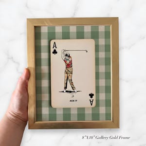 K&ouml;nnte beinhalten: Ein gerahmtes Wandbild mit einer Kreuz-Ass-Spielkarte und einer Vintage-Illustration eines Golfers, der einen Schl&auml;ger schwingt. Die Karte ist auf einem gr&uuml;n-wei&szlig;en Vichy-Karo-Hintergrund in einem 20,32 cm mal 25,4 cm gro&szlig;en Goldmetallrahmen zentriert.