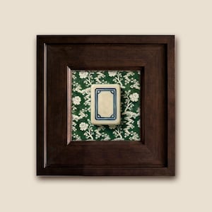 A framed white dragon Mahjong tile with a dark brown wooden frame. The tile is rectangular with a cream-colored center and a blue border. It is set against a green floral chinoiserie patterned background.
