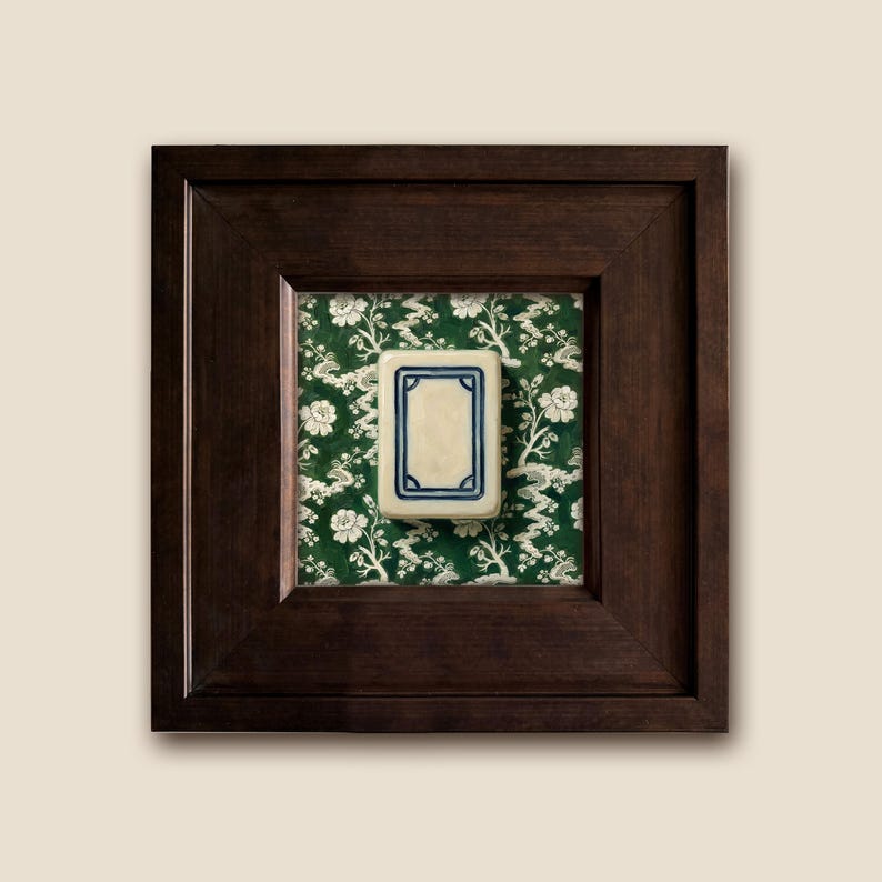 A framed white dragon Mahjong tile with a dark brown wooden frame. The tile is rectangular with a cream-colored center and a blue border. It is set against a green floral chinoiserie patterned background.