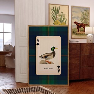 May include: Framed wall art featuring a playing card design with a duck illustration. The card is the Ace of Clubs, with the words "LUCKY DUCK" printed below the duck. The background is a green and blue plaid pattern.