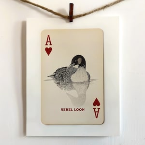 Puede incluir: Una carta de juego rectangular con una ilustraci&oacute;n de un colimbo en blanco y negro nadando en el agua. La carta muestra la letra A y un s&iacute;mbolo de coraz&oacute;n rojo en las esquinas, con el texto REBEL LOON impreso en color rojo.