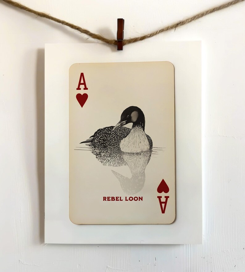 Puede incluir: Una carta de juego rectangular con una ilustraci&oacute;n de un colimbo en blanco y negro nadando en el agua. La carta muestra la letra A y un s&iacute;mbolo de coraz&oacute;n rojo en las esquinas, con el texto REBEL LOON impreso en color rojo.