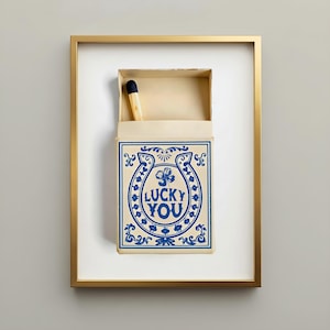 May include: Framed artwork featuring a matchbox with a single match. The matchbox is cream-colored with blue decorative elements and the words "LUCKY YOU" in a horseshoe design. The frame is gold.
