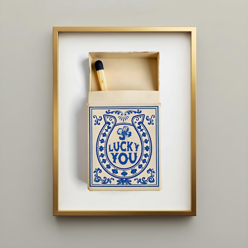 May include: Framed artwork featuring a matchbox with a single match. The matchbox is cream-colored with blue decorative elements and the words "LUCKY YOU" in a horseshoe design. The frame is gold.