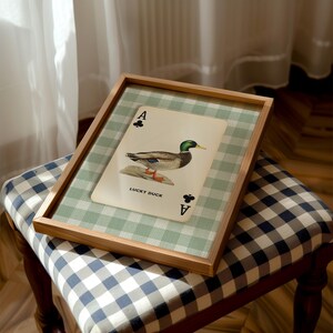 May include: A rectangular wooden frame containing a print of an Ace of Clubs playing card featuring a mallard duck illustration and the text LUCKY DUCK. The print is set against a light green and white gingham background within the frame.