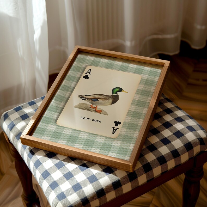May include: A rectangular wooden frame containing a print of an Ace of Clubs playing card featuring a mallard duck illustration and the text LUCKY DUCK. The print is set against a light green and white gingham background within the frame.