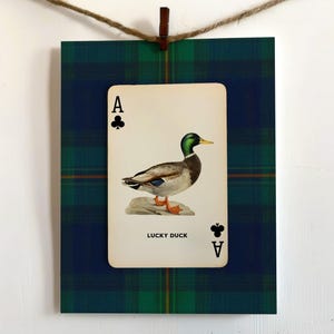 May include: A framed print featuring an Ace of Clubs playing card with a duck illustration. The card is centered on a green and blue plaid background. The text "LUCKY DUCK" is below the duck image.