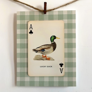 May include: A rectangular card featuring a mallard duck illustration centered on a light green and white gingham background. The card displays the text LUCKY DUCK, the letter A, and the club suit symbol in the top left and bottom right corners.