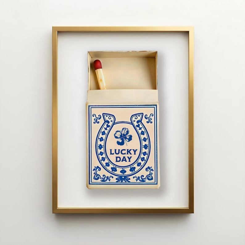 Vintage-inspired blue and white matchbox artwork featuring a single red-tipped match, horseshoe, and four-leaf clover with the phrase "Lucky Day." Framed Americana toile style wall art for western, ranch, farmhouse, or cottage interiors. Shown here in our Gallery Gold frame.