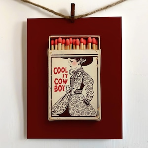 Op de afbeelding: Een ingelijste luciferdoos met een dieprode achtergrond en een vintage illustratie. De luciferdoos toont een vrouw in een patroonjurk en de tekst "COOL IT COWBOY" in het rood. De luciferdoos is open en onthult lucifers met rode uiteinden.