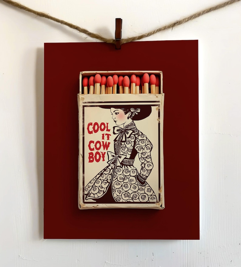 Op de afbeelding: Een ingelijste luciferdoos met een dieprode achtergrond en een vintage illustratie. De luciferdoos toont een vrouw in een patroonjurk en de tekst "COOL IT COWBOY" in het rood. De luciferdoos is open en onthult lucifers met rode uiteinden.