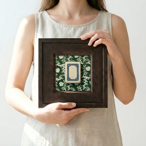 A square, dark brown wooden frame holds a print of a White Dragon Mahjong tile set against a green and white floral chinoiserie patterned fabric. The tile is centered within the frame. Shown held in a woman's hands. She is wearing a linen dress in a light-filled area.