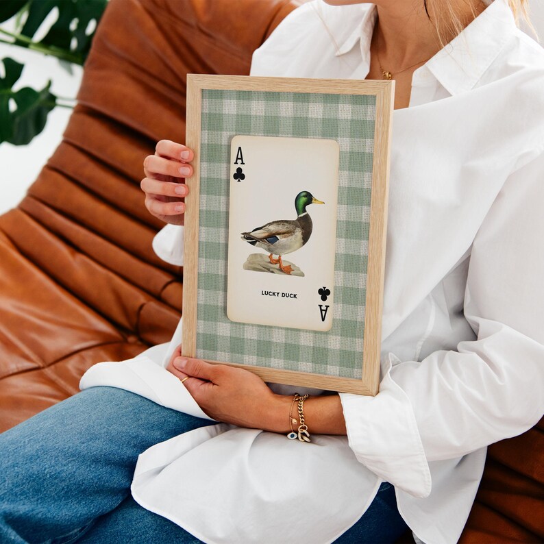 May include: A framed wall art piece featuring a playing card design with an ace of clubs symbol and a mallard duck illustration. The card includes the text LUCKY DUCK and is set against a sage green and white gingham patterned background.