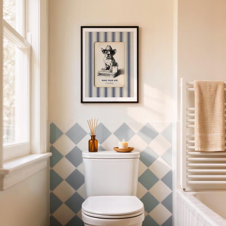 May include: A framed print featuring a bulldog wearing a cowboy hat, with the text "Mind Your Aim Cowboy." The print hangs above a toilet in a bathroom with blue and white diamond-patterned tiles. A candle and diffuser sit on the toilet tank.