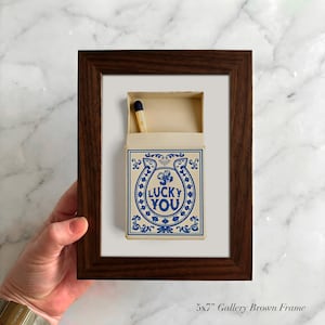 May include: A framed artwork featuring a matchbox with a blue horseshoe design and the words "LUCKY YOU." The matchbox is open, revealing a single match. The frame is a dark brown color.