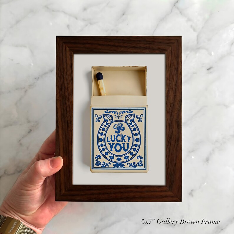 May include: A framed artwork featuring a matchbox with a blue horseshoe design and the words "LUCKY YOU." The matchbox is open, revealing a single match. The frame is a dark brown color.