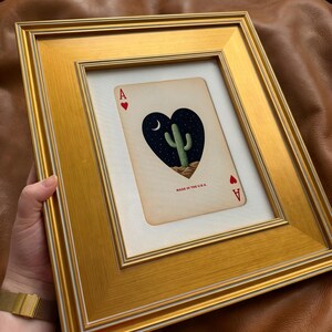 May include: Framed Ace of Hearts playing card with a gold frame. The card features a heart-shaped design with a cactus under a crescent moon and stars. The text "MADE IN THE U.S.A." is visible.