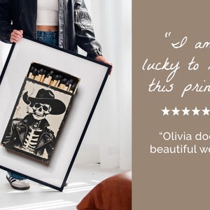 May include: A framed print of a black and white illustration of a skeleton wearing a cowboy hat and leather jacket. The skeleton is holding a cigarette and is surrounded by matches in a box. The text "I am lucky to have this print!" and "Olivia does beautiful work." is written below the image.