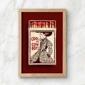 Op de afbeelding: Een ingelijst kunstwerk met een lucifersdoosontwerp. De luciferdoosillustratie toont een vrouw in een patroonjurk en de tekst "COOL IT COWBOY". De luciferkoppen zijn rood en de lijst is lichtbruin.
