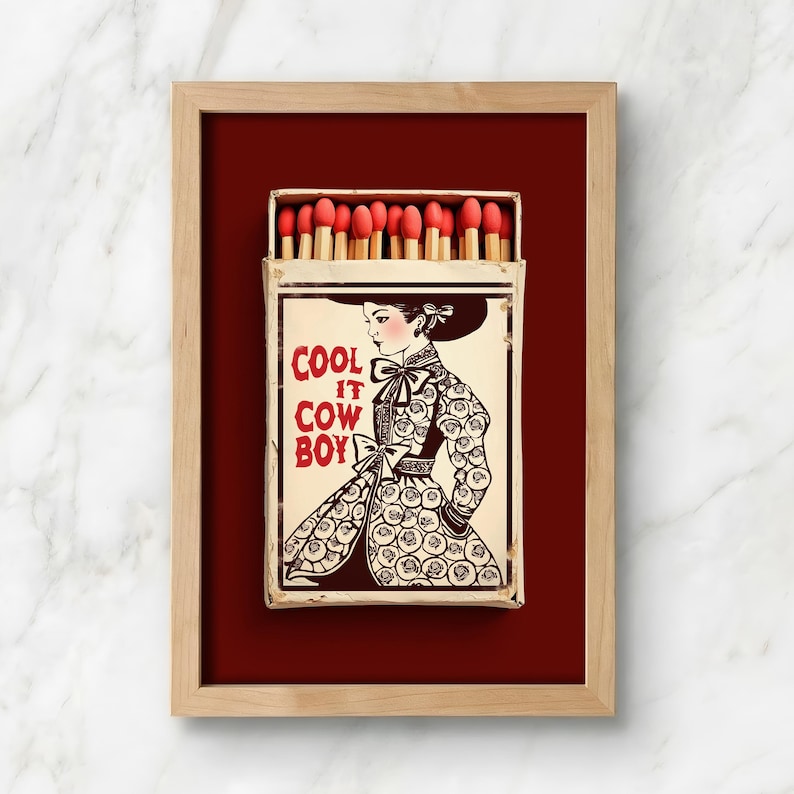 Op de afbeelding: Een ingelijst kunstwerk met een lucifersdoosontwerp. De luciferdoosillustratie toont een vrouw in een patroonjurk en de tekst "COOL IT COWBOY". De luciferkoppen zijn rood en de lijst is lichtbruin.
